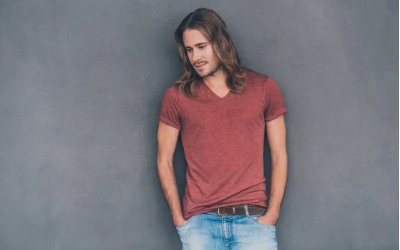 Man with a long hairstyle and his hands in his pockets leans against a wall and looks down at the ground