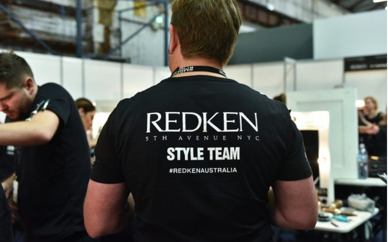A guy wearing a REDKEN style team shirt looks to his left over the person cutting hair