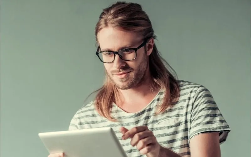 Man with half-up hair looks at a tablet wearing glasses for a bunch of long hairstyles for men
