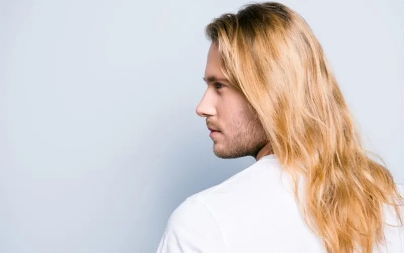 Modern Fabio long hairstyle for men showing a guy in a blue room looking left and letting his blonde dyed hair flow on a white shirt