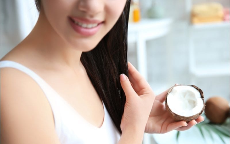 A pretty young Asian woman holding a coconut and applying the oil to her hair for a featured image for a piece on is coconut oil good for your hair
