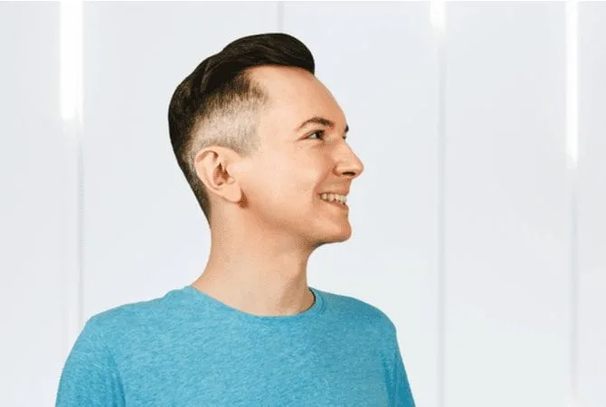Happy young man with a burst fade stands smiling in a blue shirt against a white metal backdrop