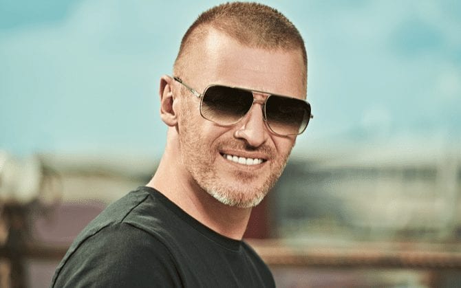 Man in sunglasses with a number three haircut stands in front of a blurry field