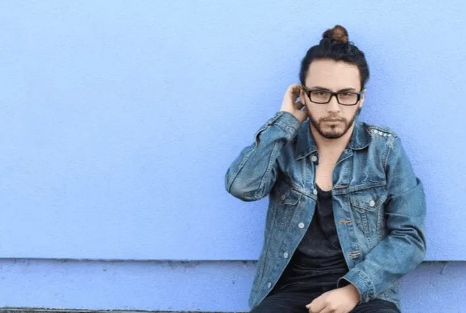 Bearded man in a jean jacket and a twisted man bun sitting against a blue wall for a piece on how to do a man bun