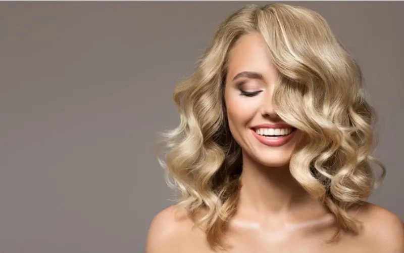 For a piece on what is hair glaze, a blonde woman smiling and closing her eyes against a grey wall with curly hair falling in front of her face