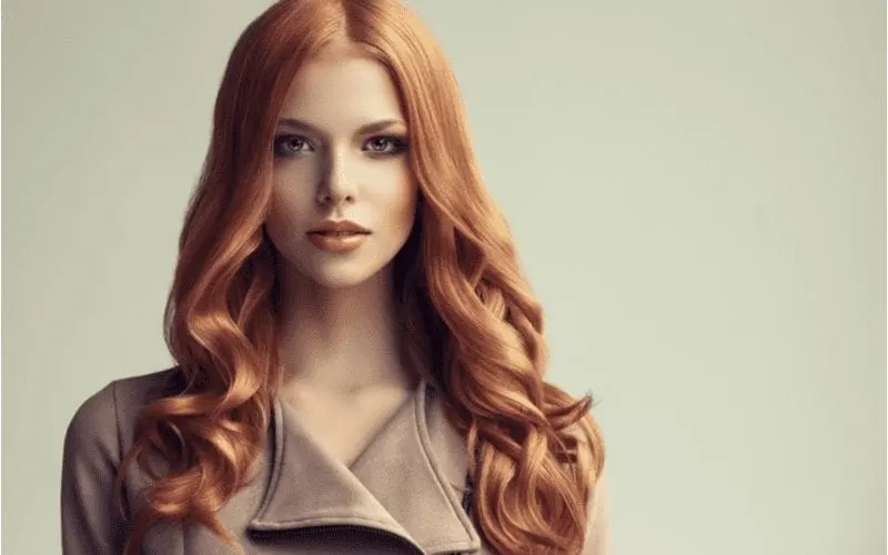 Woman with dark strawberry blonde red hair down to her shoulders looks directly at the camera and wears a trenchcoat