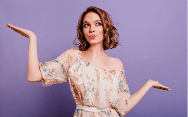 Against a purple background stands a gal with a medium-long lob haircut holding her hands in a questioning pose