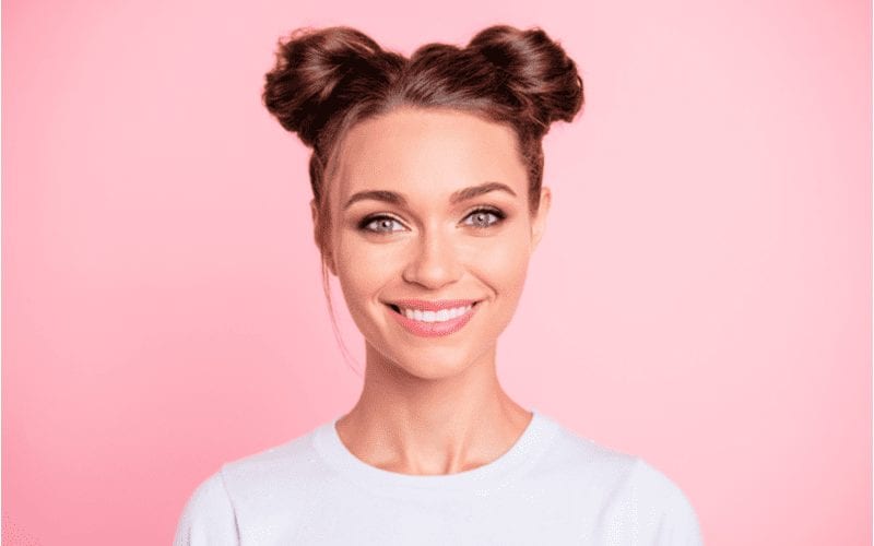 For a piece on medium-length haircuts, a woman with 90s space buns smiles in a white shirt in a pink room