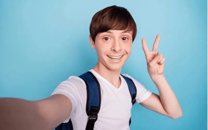 Long Side Swept Bangs on a kid with a backpack throwing up a peace sign in a blue room for a piece on boys haircuts