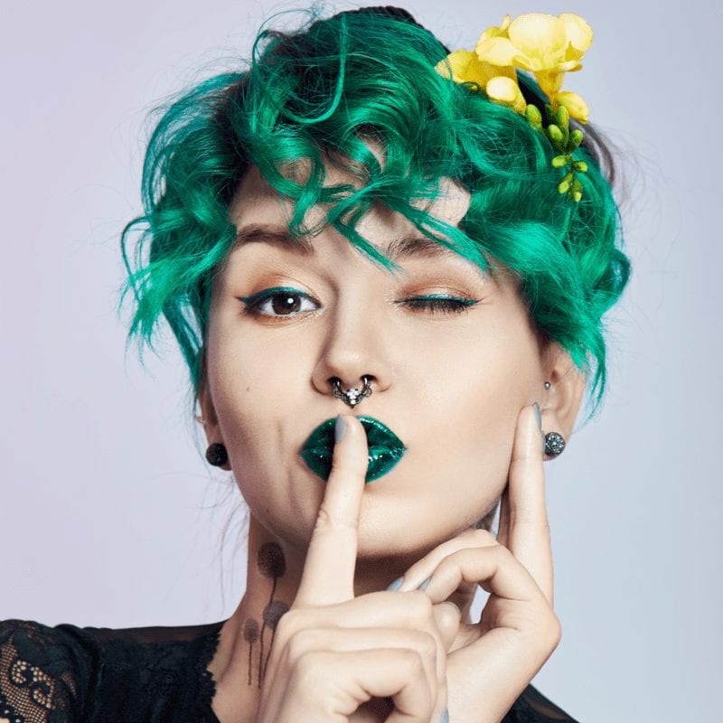 Woman with green short curly hair puts her finger up to her lips in a shush motion