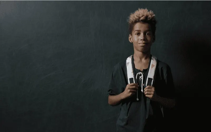 Image of an African-American boy standing with a backpack while he holds the straps in a dark room