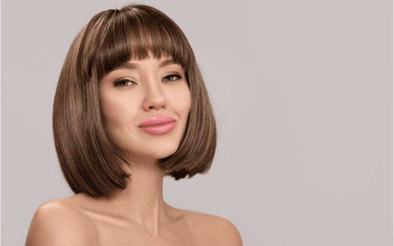 Woman with a lob haircut and no shirt that we can see grins in a studio while wearing pink lipstick