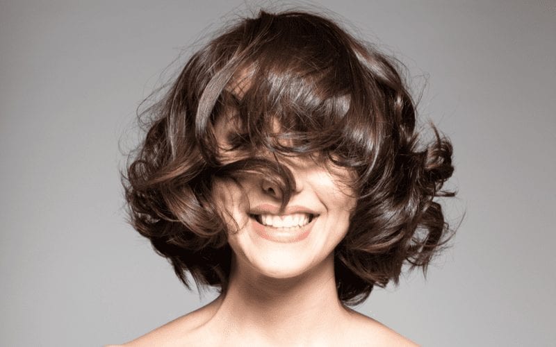 Woman who has a layered bob stands with her hair over her face and smiles big