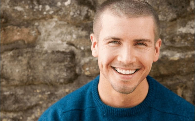 Attractive man in a blue sweater smiles standing in front of a brick wall
