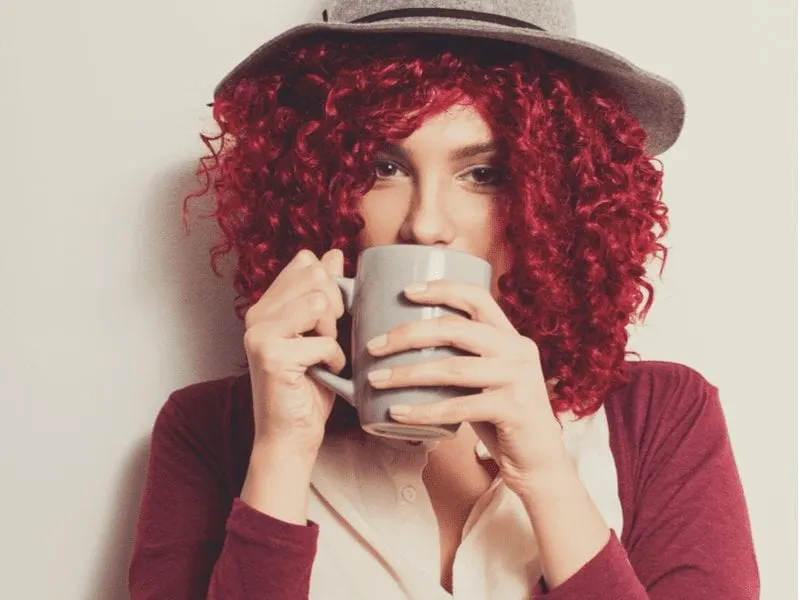 Lady with a boho hat and red hair that is curly standing and holding a coffee cup up to her mouth