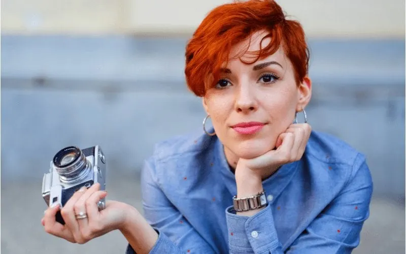 Redhead smiling and leaning forward while holding a small film camera and resting her chin on her hand