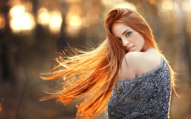 Lady with long red hair holds her waste and wears a sweater over her shoulder in the fall