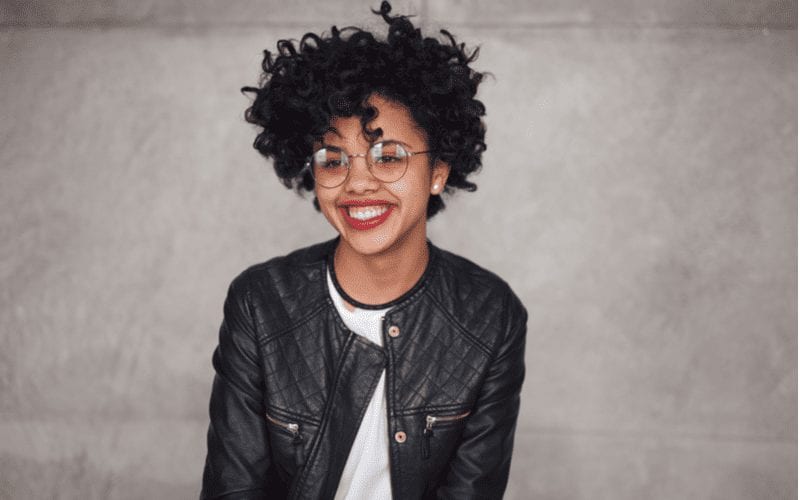 Woman with Chaotic Coils stands and smiles with hipster glasses and a black leather jacket