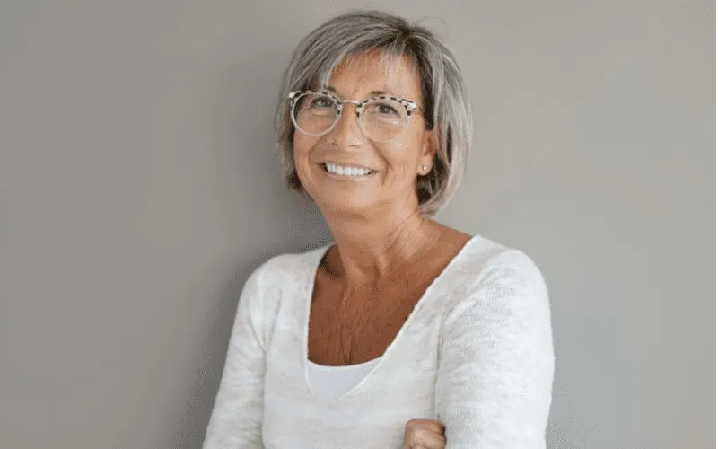 Lady with glasses meekly smiling at the camera and crossing her arms in a grey room