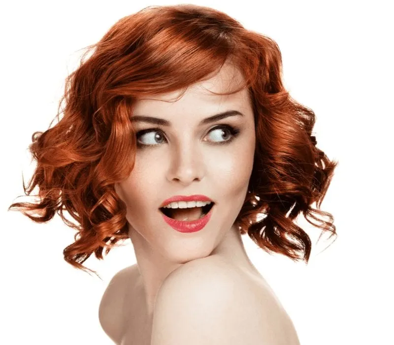 Lady smiling with red hair and standing in a white room against a white wall and looking off to her left
