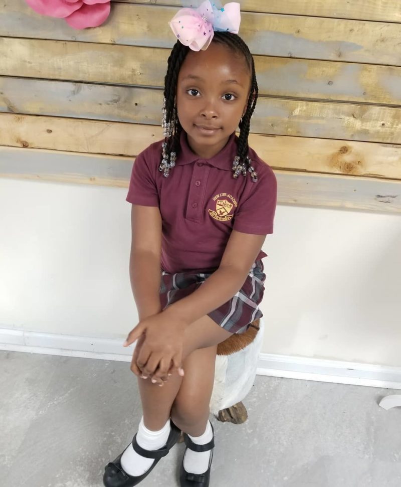 Girl in a private school outfit sits against a shiplap wall for a piece on toddlers braided hairstyles