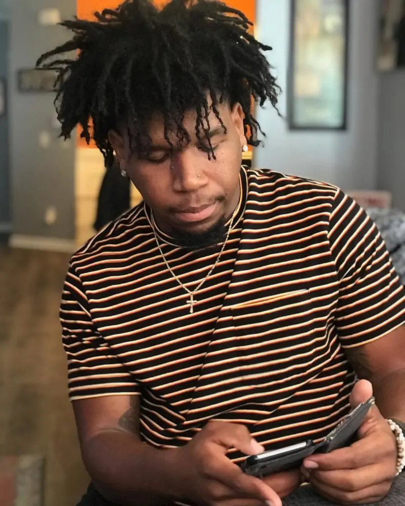 Man with freeform dreads in a yellow and black striped shirt checks his phone