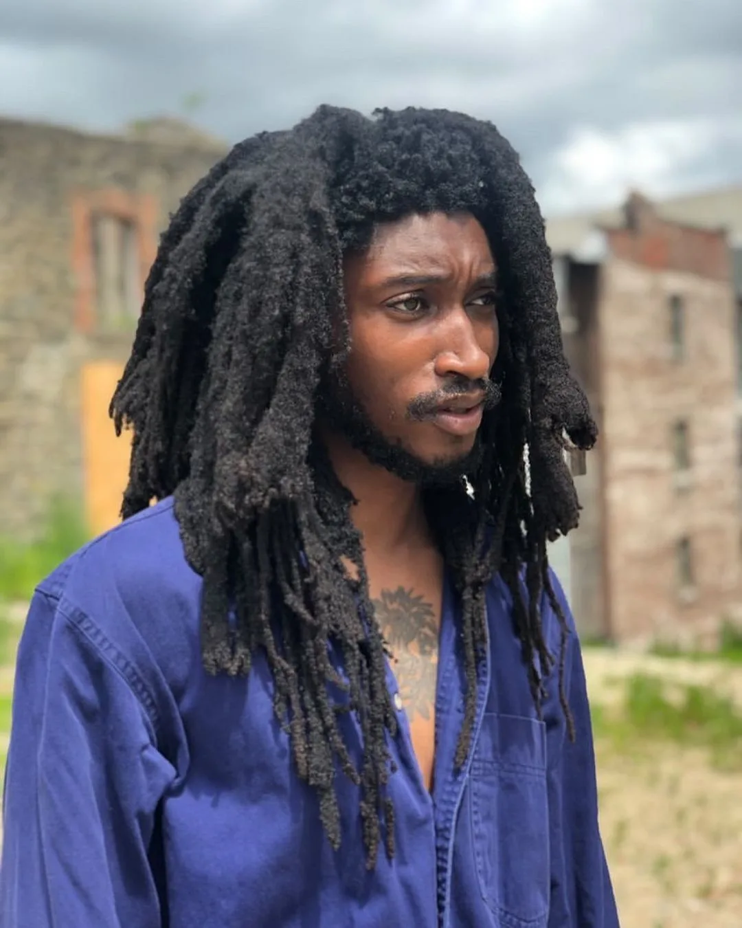 Image of a man with Congo freeform locs in a blue shirt squinting outside a brick building