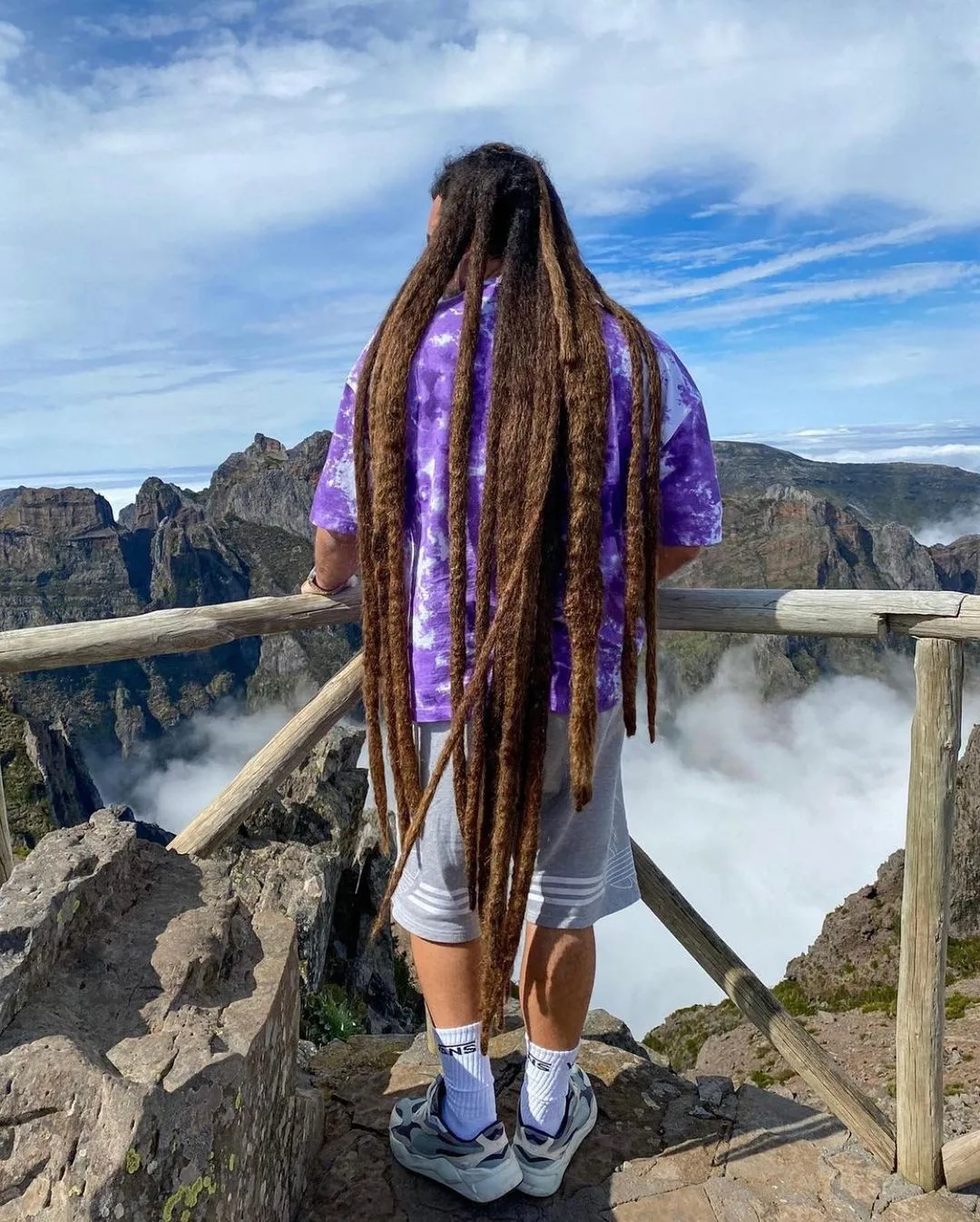 Man with insanely long freeform locs standing in front of a fence that overlooks a volcano