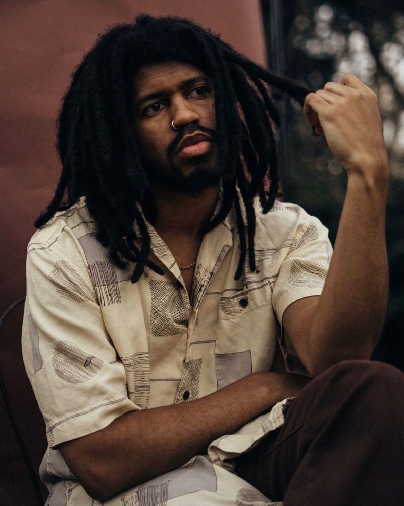 Man with thick freeform locs holds one of the strands in his left hand and contemplates something while sitting