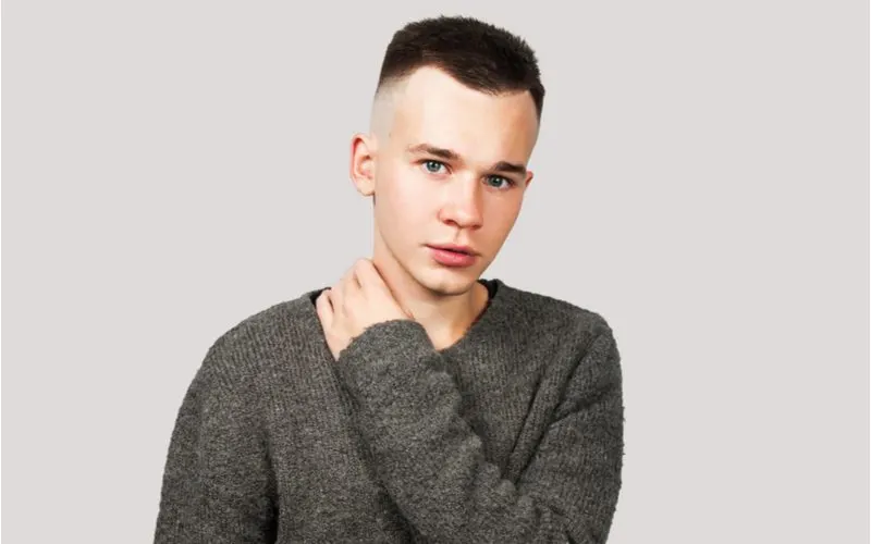 Young man in an oversized grey wool sweater holds his neck while wearing an undercut fade in a studio