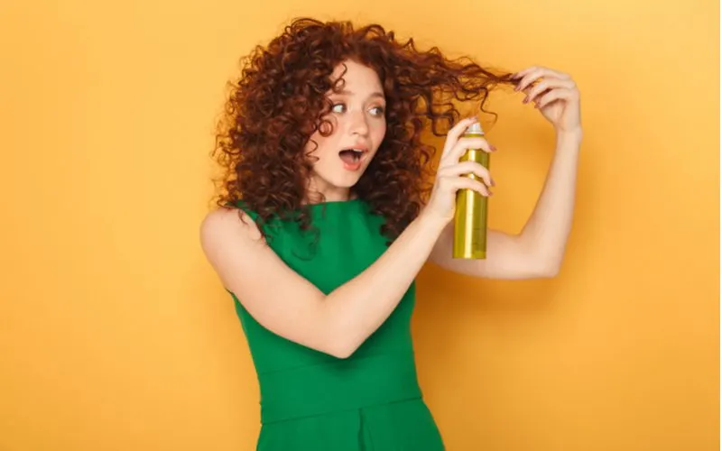 Woman spraying her curly red hair with a hair thickening product and standing in a green dress in an orange room