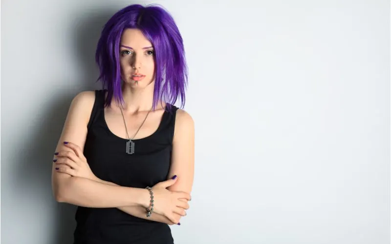 Portrait of a girl with bright purple emo hair crossing her arms in a black shirt and leaning against a wall