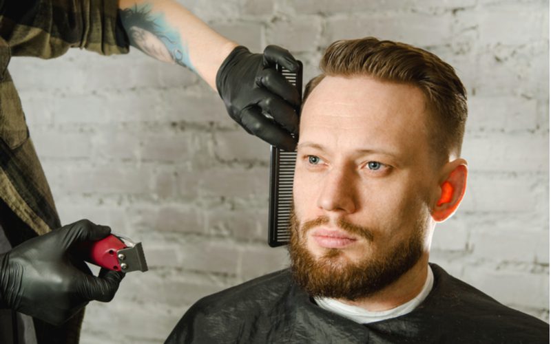 man wears a high and tight undercut fade while sitting without smiling in a barber chair and getting groomed by a guy in black gloves