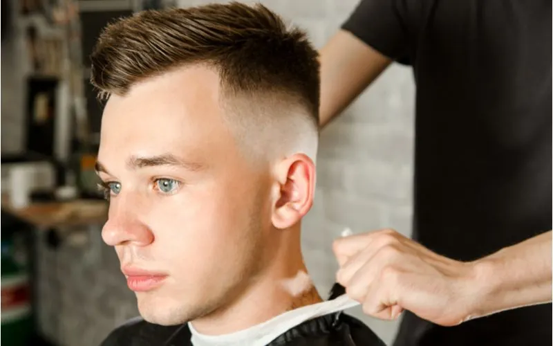 Man with a wavy side parted undercut fade haircut has his cape unbuttoned in a barber's chair and looks ahead without smiling
