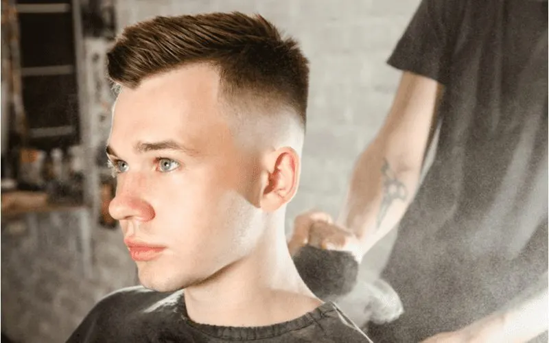 The hairdresser cleans the neck of a young guy with a brush with talc after a haircut against a brick wall background