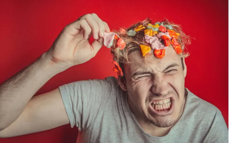 Man with a bunch of gum in his hair winces as he pulls a piece out while standing in a red room