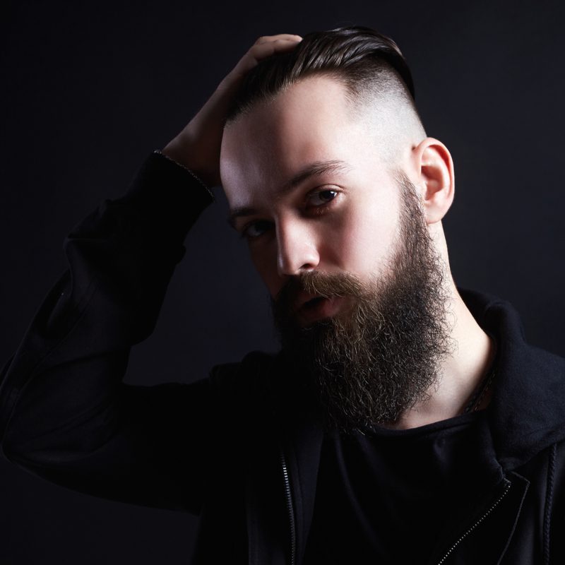 Man with a bearded hipster undercut fade grabs his head and wears black clothes in a dark room