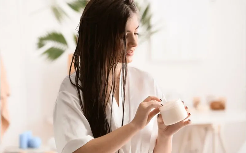 Woman in a white robe with beautiful long hair learning how to clarify hair by reading these steps while she puts her hand into a tub of hair mask