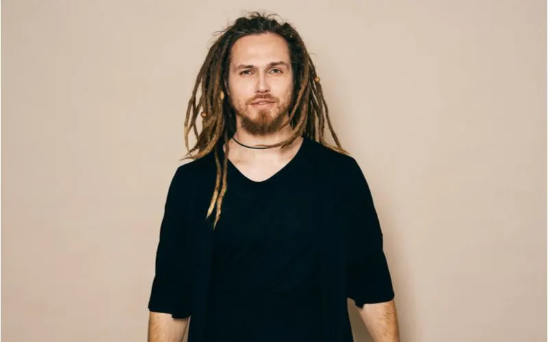 As an image for a piece on how to freeform dreads, a man with such a style in a black shirt stands in a tan studio room