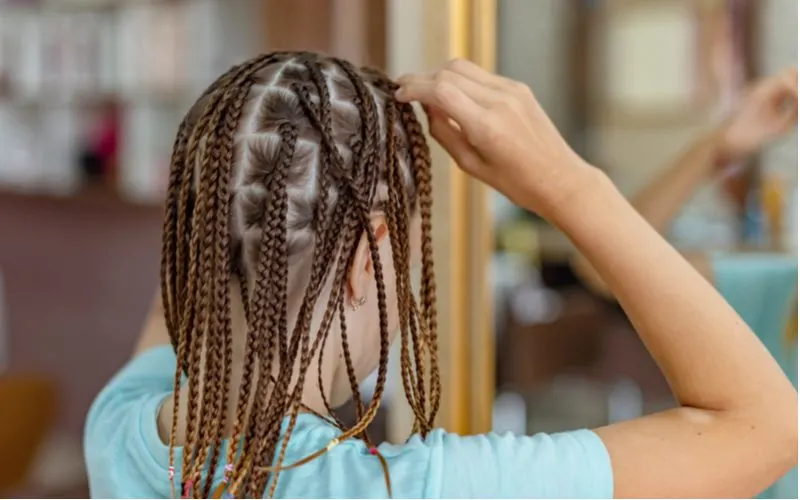 A woman wearing box braids holds the front of her head while looking in a mirror after doing box braids on herself