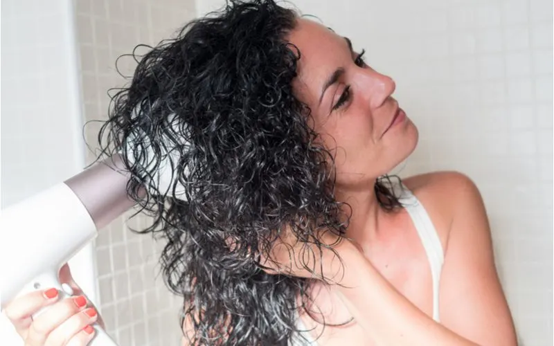 Image of a woman showing us how to diffuse hair and using a blow dryer with such an attachment while standing in a white bra while smiling