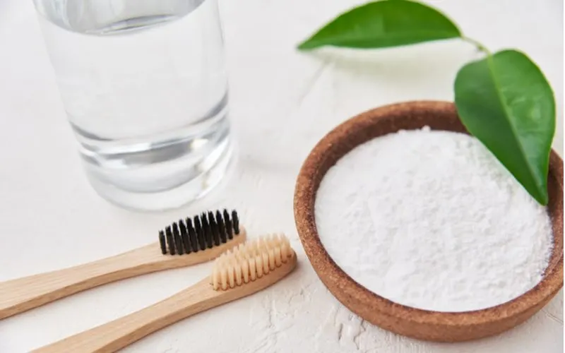 Image of two wooden tooth brushes sitting next to baking soda and leaves and water