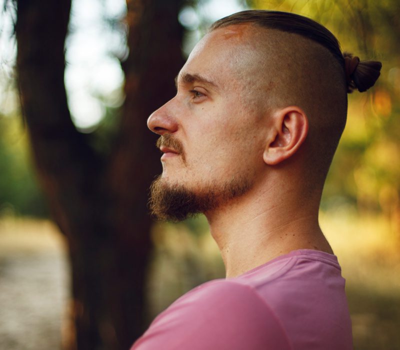 Hipster man with an undercut fade with bun stands in the forest crossing his arms and holding his goateed chin in the air