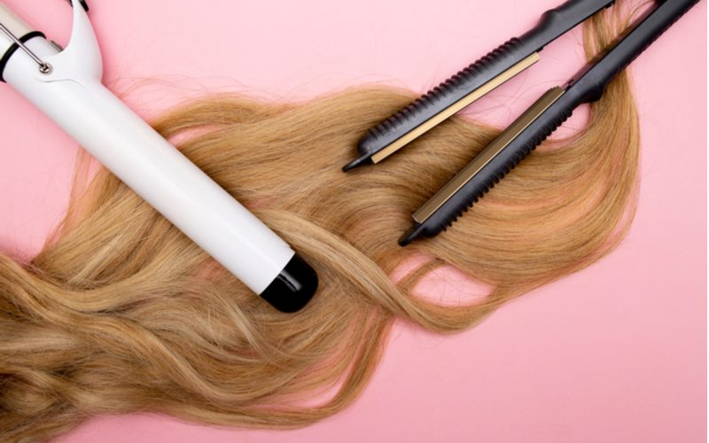 To help illustrate how to get thicker hair naturally, a couple hot tools to avoid using sitting on top of blonde hair on a pink background