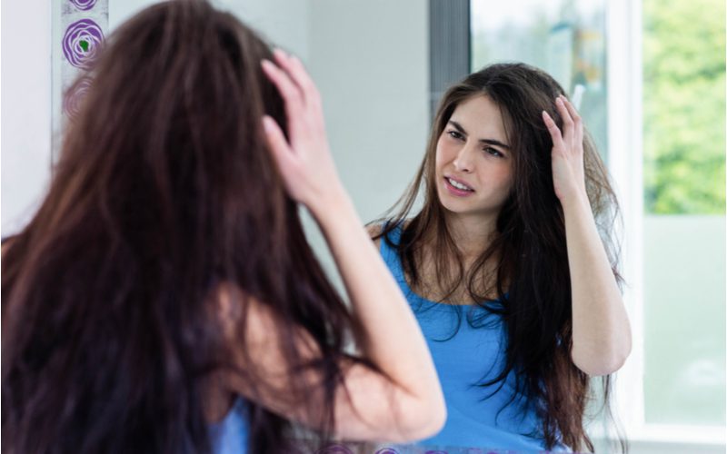 Brunette looking in the mirror and showing visible displeasure because she doesn't know how to get rid of frizzy hair and holding the right side of her head