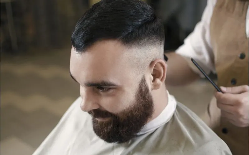 Man wears a neat undercut fade while sitting smiling in a barber's chair and wearing a very thick beard