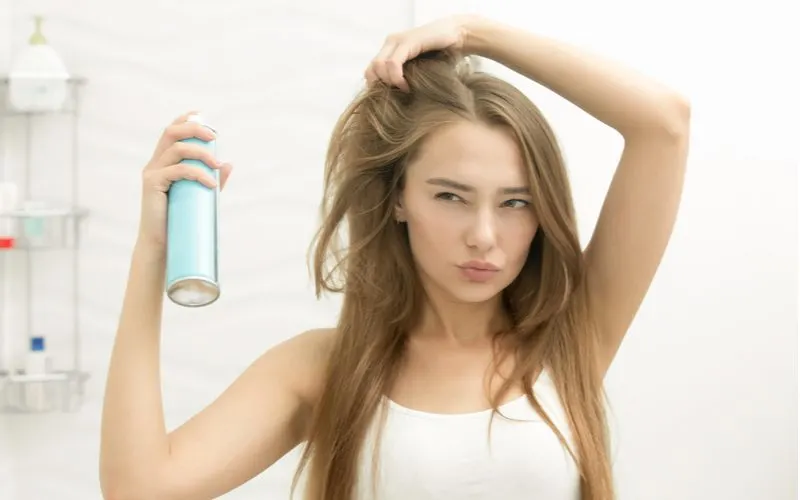 Girl in white cami using the best setting spray on her brown hair while holding her right curtain bangs up