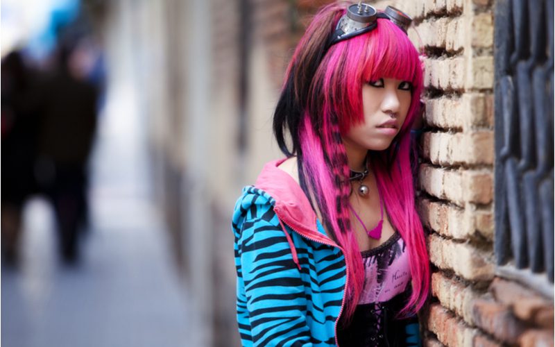 Pink haired asian woman with emo hair wearing a blue zebra hoodie and a tight spandex top standing against a brick wall