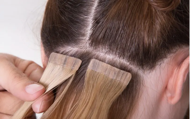 To illustrate how long extensions last, a woman getting taped extensions on the back of her head