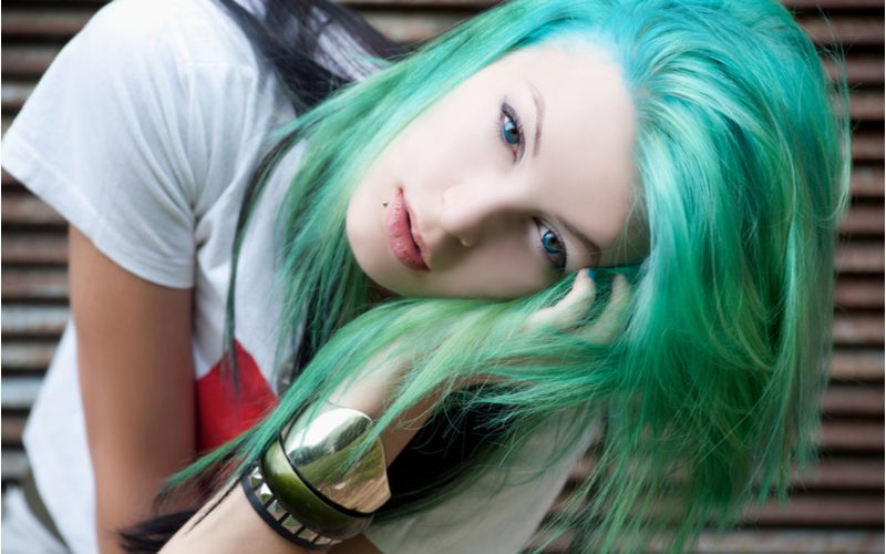 Girl in a white shirt with bright green balayage green hair wearing metal bracelets holding her face